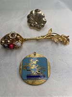 Photo showing three vintage jewelry items: a silver floral brooch, a gold-tone embellished spoon brooch with gemstones, and a square cloisonne pendant with blue and gold floral design.