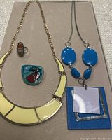 Photo of four pieces of vintage enamel jewelry laid flat on a beige background including two necklaces and two rings.