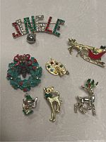 Photo showing 8 Christmas-themed vintage brooches and pins including 'Jingle' rhinestone word pin, wreath, elf head, cat, sleigh, reindeer, and palette.