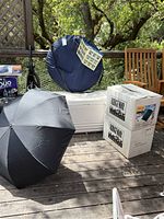 Full view of camping equipment items including two Ridgeway packaged sleeping bags, large white Igloo cooler, black folding umbrella, and circular blue bag likely for the tent.
