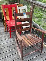 Seven wooden chairs on a wooden deck including a worn vintage dark-stained rocking chair and six upright chairs in various colors – red, white, natural wood.