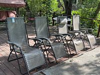 Four metal reclining patio chairs arranged side by side on a wooden deck with visible sunlight and shadow. Chairs have vinyl mesh seats and plastic armrests.