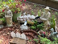 Two cement standing figures (girl with basket, boy with hat), composite sleeping angel figure, vintage flower pot, slate, quartz rock, and plants in garden bed.