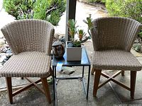Two wooden outdoor chairs with vinyl woven seats and backs flanking a black metal and glass side table, with a plant in pot and carved monkey figurine.