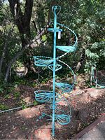 Full view of tall blue painted iron plant stand with eight circular shelves arranged vertically outdoors among trees and foliage.