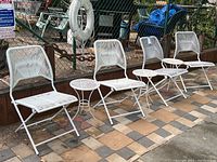 Four white metal folding chairs with vinyl mesh backs and seats, lined up outdoors on a patio with tiled flooring and chain-link fence background.