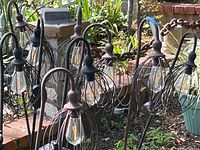 Ten black metal cage style solar garden lights installed in garden beds, showing their bulb-shaped LED lights, metal stake mounts, and attached solar panels