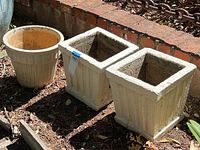 Three garden planter pots placed outdoors: two white square cement pots with ribbed design and one round ceramic pot with ribbed vertical pattern.