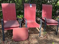 Three red outdoor chairs with steel frames and nylon mesh backs and seats, outdoors on soil ground. One chair's seat cushion is removed and placed in front, showing red cushioned pad.