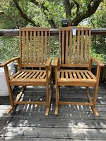 Front view of two wooden rocking chairs showing width and vertical slat design on seat and backrest, with armrests and rockers on deck flooring outdoors.