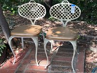 Front view of two Mid Century Modern cast iron chairs with off-white lattice oval backs and tan vinyl cushions, outdoor setting on brick and dirt ground.
