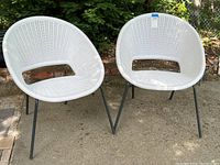 Two white Nautica outdoor satellite chairs side by side on concrete ground outdoors with greenery in background. Chairs have black metal legs and white faux mesh seating and backs.