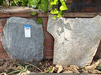 Two slate stones propped against a brick wall with foliage above and dried leaves below, showing relative size and texture.