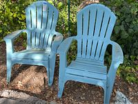 Two blue plastic high back chairs shown outdoors on dirt ground with foliage backdrop. Chairs display signs of use and outdoor exposure.