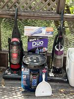 Wide shot of all vacuum and cleaning equipment including the upright vacuums, boxed Shark and Black & Decker handheld vacuums, Roomba robot vacuum, Bissell ProHeat spot cleaner, and a white dustpan.