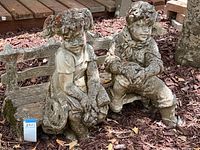 Full view of vintage cement sculpture showing girl and boy seated on decorative bench, with ground mulch and wooden deck background.