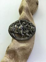 Front of the oval silverplated brooch featuring cherub and floral cutout design fixed on a wood piece.