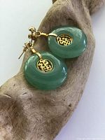 Close-up of green jade earrings with gold plated Chinese character medallions, displayed on a carved wooden object.