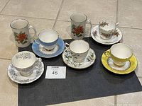 Photo showing all 12 pieces including various teacups, saucers, and mugs with multiple floral and scenic designs on display on the floor.