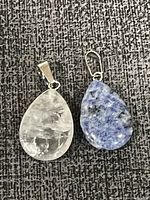 Two crystal pendants laid on textured fabric, showing clear and blue stones with silver-tone bails.