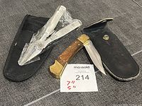 Two utility tools placed on a grey fabric surface: a stainless steel multi-tool partially wrapped in plastic next to its black nylon sheath, and a folding knife with a wood handle and brass bolsters accompanied by a black faux leather sheath.