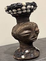 Front side of carved statue piece showing face and cowrie shell adornments on head and base.