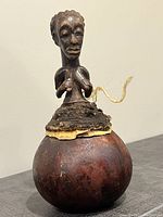 Front view of antique carved wooden rattle with attached human figure, showing detailed face, hair and rounded base with visible wear and surface markings.