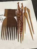 Photo showing two carved wooden combs and six carved hair sticks with giraffe and plain designs laid out on a white surface with a MaxSold tag.
