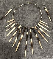 Front view of necklace and matching earrings lying flat on grey fabric surface. Necklace features multiple elongated tapered pendants in dark brown and cream gradient colors, arranged by size and paired with smaller beads. Earrings match with single tapered pendants of similar color gradient.