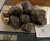 Photo of multiple raw amber stones placed on a scale showing total weight of 652 grams. Stones are rough, dark brown to black in color with natural surfaces.
