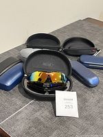 Lot of sunglasses and multiple sunglasses cases arranged on a table. Includes one open black case with reflective sunglasses inside and six additional hard sunglasses cases around it.