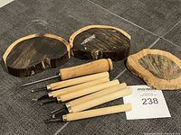 Photo showing five wood carving tools with wooden handles and three dark wood pieces, likely ebony, on a dark tile floor.