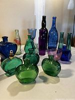 View of 16 glass bottles and vases in colors green, blue, and purple arranged on a white surface. Shows varying sizes, shapes, and textures including heart-shaped, rounded, and tall slender bottles.