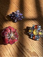 Three women's decorative pins laid on a wooden surface showing their colors and shapes: deep pink flower, sparkly pale lavender pink stone flower, and multicolor stone round pin.