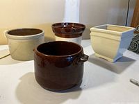 Four ceramic planter pots in brown and cream shades arranged together on a white surface; front dark brown pot has small handles