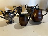 Photo of five assorted antique metal chalices, pitchers and a mug on flat surface showing wear and tarnish.