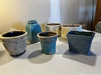 Photo showing six ceramic pots of varying shapes and sizes in cream, sea-foam green, and aqua colors arranged on a table.