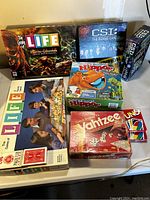 Photo of assorted board games and card decks arranged on a table showing titles including The Game of Life, The Game of Life Pirates of the Caribbean, Yahtzee, Hungry Hungry Hippos, CSI Board Game and CSI Crime Game.