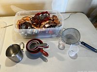 Overview showing plastic container with very assorted metal and plastic cookie cutters and small cake molds, nested measuring cups, two small liquid measuring shot glasses, a metal sifter, and small metal pitcher.