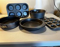 Various used cake and cupcake pans on a light-colored surface including 2 large round cake pans, 1 large cupcake pan with 6 cavities, 1 mini cupcake pan with 12 cavities, 1 springform pan with latch, and 1 bundt pan with removable bottom.