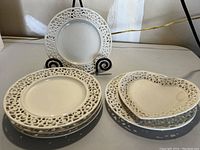 Six cream-colored dessert plates with lace edge pattern, five round and one heart-shaped, displayed on and near a black metal stand.