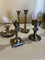 Five assorted metal candlesticks displayed on a surface, showing variety in height, base design, and finish including one labeled solid brass.