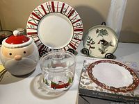 Miscellaneous Christmas-themed dishes including large platter, small plate with bird design, Santa Claus cookie jar, and plastic container with truck design, including boxed red Fine Stoneware vegetable platter.