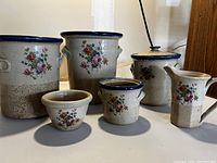 Full view of the Monroe Saltworks set showing four canisters, creamer, sugar dish, and a small item with floral decoration on brown speckled ceramic with blue rims.