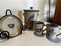 Photo showing the set of Monroe Saltworks ceramic items including canister with lid, two mugs, and a plate on a stand, all decorated with bird images on a brown speckled background.