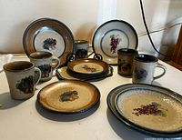 View of all mugs and plates showing the fruit and floral patterns and color scheme with brown background and green and blue rims.
