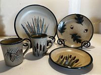 Set of three nature pattern plates and two mugs arranged on table. Plates show cattail design, acorn and leaves. Mugs show cattail and pinecone designs.