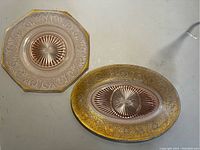 Two rose-tinted glass plates: one octagonal and one long oval, both with gold-colored etched floral designs and visible wear to gold detailing.