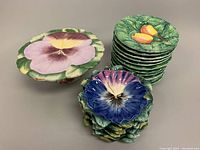 Twelve hand painted Majolica plates stacked: six with peach fruit design on green leaf background and six with purple pansy floral design. Pedestal cake plate visible.