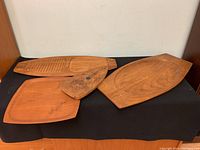 Four teak serving trays arranged on a black surface showing different shapes and sizes, highlighting medium brown wood tone and carved details.